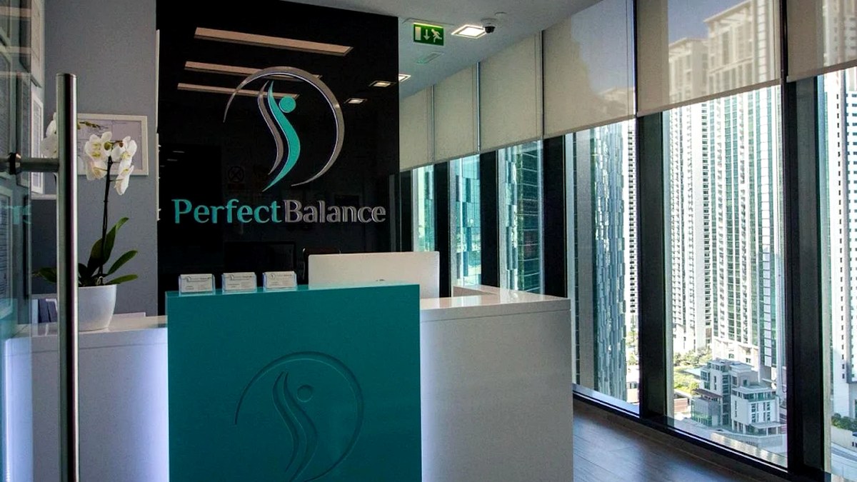 Reception desk of a modern office with a turquoise front panel and the 'Perfect Balance' logo on a dark wall behind it, near tall windows overlooking a city skyline